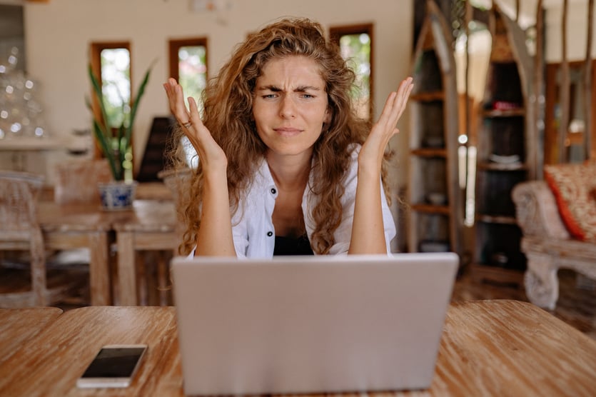 Kvinde sidder ved bord med bærbar computer og ser undrende ud med hænderne oppe